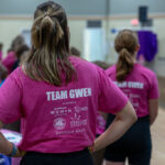 Two women wearing matching “TEAM GWEN” pink t-shirts stand in a room with decorated tables, watching someone speak on a stage in the background. The focus is on the text and sponsors on the back of one shirt.