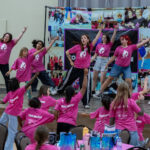 A group of girls in matching pink shirts dance energetically on stage, while more girls in similar shirts follow along in the audience. The room is decorated with purple and pink, and a colorful photo backdrop is behind the performers.