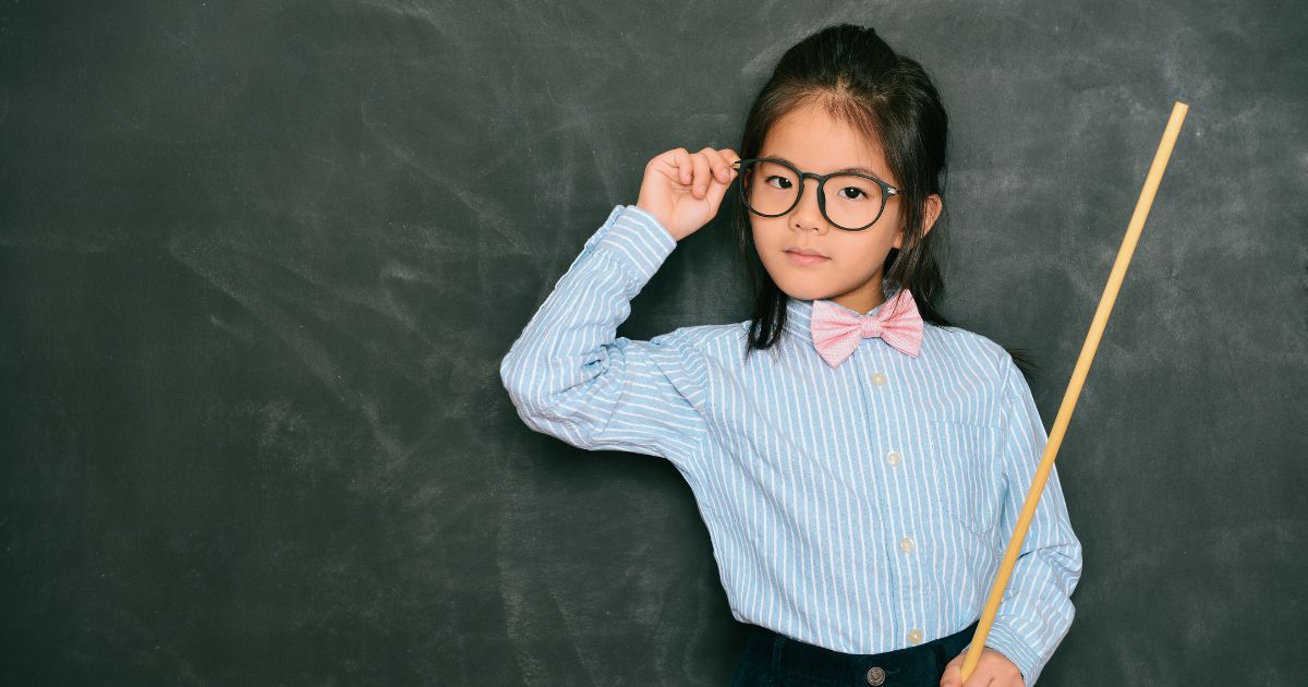 Young girl standing confidently in front of a chalkboard wearing glasses and a bow tie, symbolizing leadership and mindset coaching for girls in Alberta after her parents enrolled her to Girls On Fire conference.