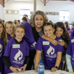 A smiling woman stands with six girls wearing matching purple Girls on Fire 2025 shirts at a busy indoor event, all looking at the camera. The girls have name tags and appear happy and excited.